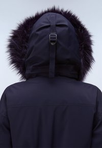 Person som bär en svart vinterjacka med huva som har mörk pälsdetalj och en justerbar rem på baksidan.