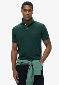 Polo shirt verde scuro con colletto, due bottoni e logo. Maglione verde chiaro annodato in vita, jeans in denim e un orologio.