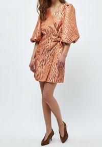 Korts korgklänning i orange med tigerstripemönster och puffärmar, gjord av ett glansigt, slätt tyg, knuten i midjan. Bruska högklackade skor.
