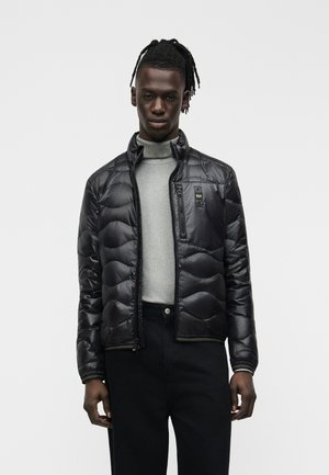 Veste noire matelassée fine portée par-dessus un col roulé gris clair et un pantalon noir sur une personne debout devant un fond uni.