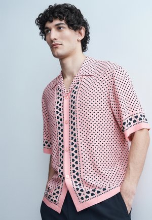 Jeune homme aux cheveux bouclés portant une chemise rose à manches courtes à boutons, ornée de petits et grands cœurs noirs, mains dans les poches.