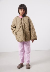 Jeune enfant aux cheveux foncés portant une veste beige matelassée trop grande, un pantalon en velours côtelé rose et des chaussures marron sans lacets, se tenant devant un fond blanc.