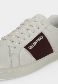 Baskets blanches avec lacets blancs et un panneau latéral rouge foncé portant le nom de la marque Valentino en lettres blanches.