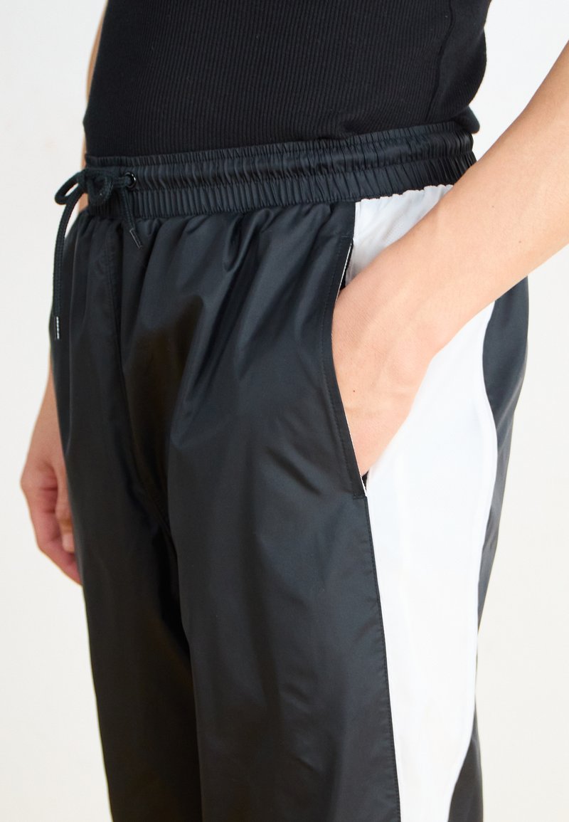 Pantalons de sport noir et blanc avec une taille élastique, poches latérales et finition brillante, présentant un panneau blanc contrastant sur le côté.