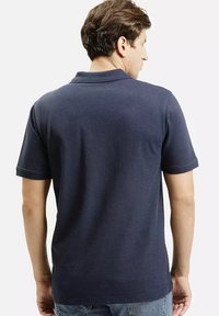 Granatowa koszulka polo o klasycznym kroju, krótkich rękawach i gładkiej fakturze. Posiada kołnierzyk zapinany na guzik oraz subtelne detale przeszyć.