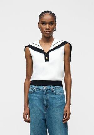 Moteris su pintinėmis šviesiais plaukais, vilkinti baltą berankovį viršų su juodu jūreivio apykakliu ir mėlynais aukšto liemens džinsais, stovinti prie balto fono.