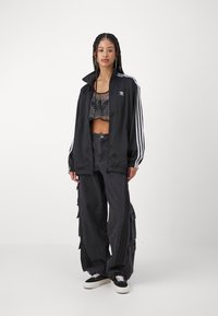 Chaqueta de chándal negra con rayas blancas, top de malla recortado, pantalones anchos negros con inserciones laterales y zapatillas negras con suelas blancas.
