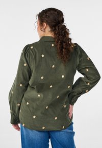 Olivgrünes Cordhemd mit gepufften Ärmeln, dekorativen bestickten Blumen und einem Hemdkragen. Getragen zu blauen Jeanshosen.