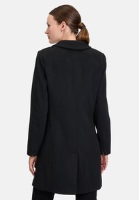 Schwarzer Wollmantel mit strukturierter Silhouette, Reverskragen und langen Ärmeln. Saubere Linien und dezente Nähte im Rückenbereich.