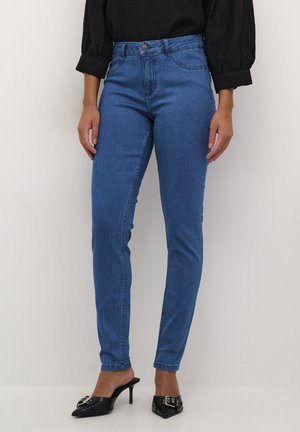 Woman wearing high-waisted blue skinny jeans, black long-sleeve blouse with puff sleeves, and black heeled mules with buckle detail.