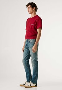 Rød bomulls-T-skjorte med brystlomme, kombinert med lyseblå slim-fit jeans og hvite joggesko med svarte detaljer. Sideprofil.