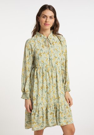 Vrouw met lang bruin haar die een lichtgroene bloemenjurk met lange mouwen en een gerimpelde kraag draagt, staand voor een witte achtergrond.