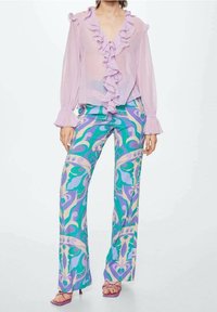 Jasnofioletowa bluzka z falbanami przy dekolcie i mankietach, połączona z niebieskimi i zielonymi spodniami w zegary. Sandały mają blokowy obcas.