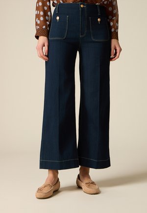 Jean bleu marine à jambes larges avec poches avant boutonnées, porté avec des mocassins couleur caramel et un chemisier marron à pois.