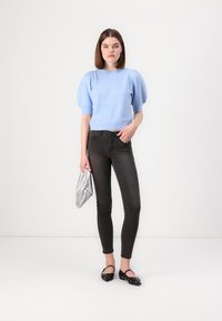 Maglione azzurro chiaro con maniche a sbuffo abbinato a jeans skinny neri e scarpe nere flat. Il modello tiene in mano una borsa a tracolla argento. Sfondo semplice.