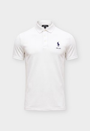 Homme debout portant un polo blanc, un short noir avec ceinture, des chaussettes blanches et des baskets blanches sur un fond clair uni.
