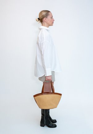 Femme en chemise blanche oversize, short beige et bottes noires tenant un grand sac à main beige et marron debout de profil sur fond blanc.