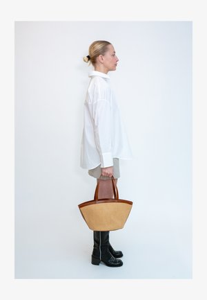 Femme en chemise blanche oversize, short beige et bottes noires tenant un grand sac à main beige et marron debout de profil sur fond blanc.
