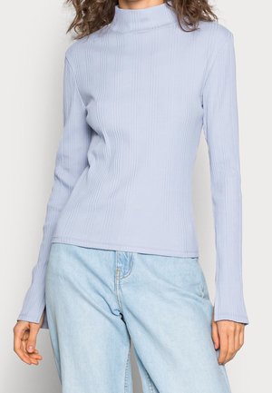 Haut à col roulé bleu clair à manches longues côtelées, porté avec un jean en denim clair, modèle montré de l'épaule aux hanches.