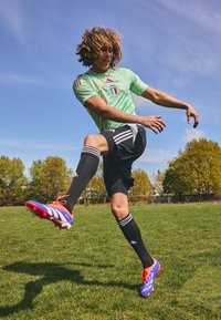 Voetballer in een lichtgroen shirt met een logo en zwarte shorts. Draagt meerkleurige voetbalschoenen, zwarte sokken met witte strepen, en is in beweging op gras.