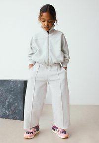 Sweat à capuche zippé gris clair avec des poignets côtelés, associé à un pantalon de survêtement gris ample et des baskets multicolores avec des accents de motif léopard.