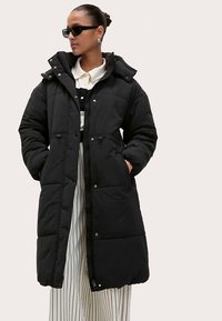 Abrigo largo negro acolchado con capucha, con cierres de botones a presión, dos bolsillos frontales y textura acolchada. Estilizado con un conjunto a rayas.