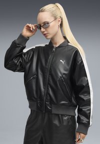 Giubbotto bomber in pelle nera con polsini a coste grigi, tasche con zip e strisce laterali bianche. Presenta un logo Puma sul petto.
