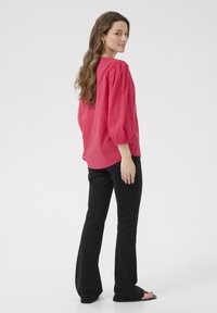 Blouse rose texturée avec des manches bouffantes, associée à un pantalon noir évasé. La blouse présente un col rond et des motifs subtils.