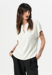 Witte blouse met korte mouwen en een sleutelgat-halslijn met subtiele zilveren accenten. Zachte, gladde stof valt iets uit bij de zoom.