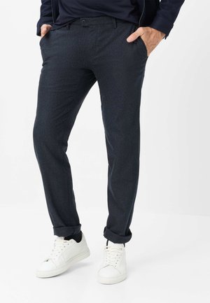Mand iført mørkegrå slim fit bukser med manchetter og hvide sneakers, hænderne i lommerne, står mod en hvid baggrund.