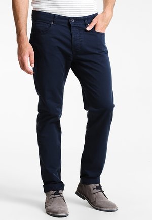 Mann trägt marineblaue Slim-Fit-Hose mit umgeschlagenen Säumen und graue Wildleder-Schnürschuhe, eine Hand in der Tasche.