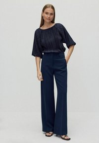 Mørkeblå plisseret bluse med brede ærmer, parret med højlivede, lige bukser. Outfittet har minimalistisk design og glatte teksturer.