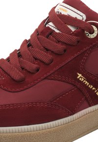 Gros plan d'une basket en daim et tissu rouge avec semelle en caoutchouc texturée, lacets rouges et logo doré "Tamaris" sur le côté.
