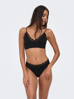 Młoda kobieta pozująca w czarnym biustonoszu z koronkowym wykończeniem i dopasowanych, wysokich majtkach na prostym, jasnym tle.