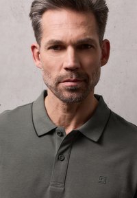 Homme d'âge moyen avec des cheveux courts et une barbe, portant un polo gris foncé, sur un fond uni gris clair.