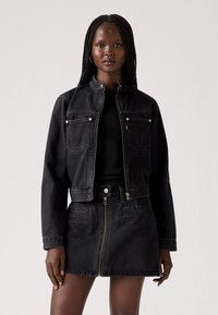 Chaqueta de mezclilla negra con cuello alto y dos bolsillos en el pecho, combinada con una falda de mezclilla negra a juego con cremallera frontal.