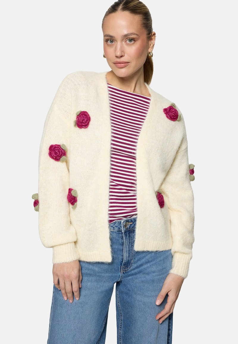 Femme portant un gilet crème orné d'appliqués de roses roses sur une chemise rayée rouge et blanche et un jean bleu, debout devant un fond uni.