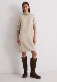 Strickkleid in Creme mit hohem Kragen, langen Ärmeln und lockerem Schnitt. Kombiniert mit dunkelbraunen knielangen Wildlederstiefeln.