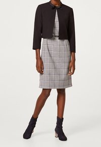 Veste noire courte avec col et manches trois-quarts portée sur une robe à carreaux grise. Bottines bleu marine à cheville avec texture plissée.