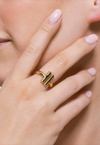 Anello in oro con un design moderno caratterizzato da una forma a croce. Texture liscia con finitura lucida, indossato su una mano con unghie morbide e di colore chiaro.