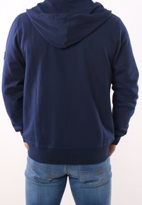 Granatowy hoodie z miękkiego materiału, z kapturem na sznurku, kieszenią kangurką i ściągaczami u rękawów, w połączeniu z jasnoniebieskimi dżinsami.