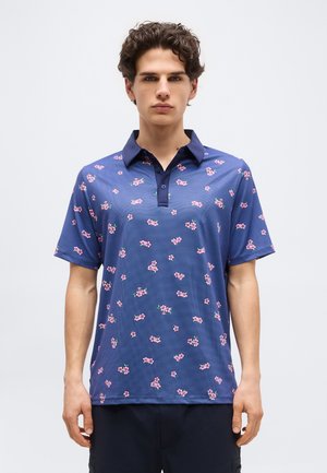 Jeune homme aux cheveux bouclés et foncés portant un polo bleu marine à manches courtes avec un motif de petites fleurs roses, debout devant un fond clair uni.