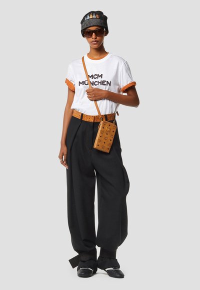 Personne portant un t-shirt blanc « MCM München », un pantalon large noir, un bonnet rayé, des lunettes de soleil, une ceinture marron et une pochette portée en bandoulière.