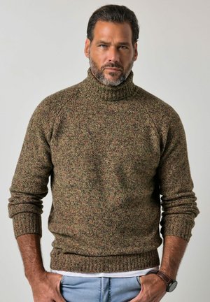Homme aux cheveux courts et foncés avec une barbe, portant un pull à col roulé tricoté marron et vert et un jean bleu clair, debout les mains dans les poches.