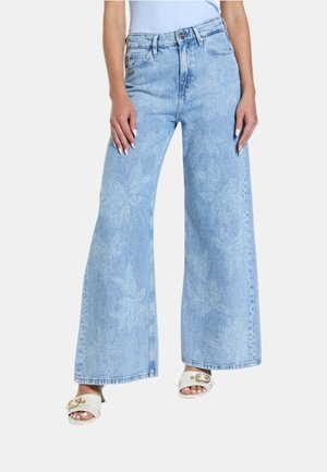 Jeans azzurri chiari a gamba larga con sottili motivi floreali, indossati con sandali bianchi con tacco e punta aperta e top smanicato infilato dentro.