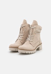 Un paio di stivaletti alla caviglia in suede beige con lacci, suole spesse e cerniere laterali su sfondo bianco.