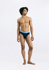 Námornícke modré plavky s ľahko modrým pásom označeným "TOMMY HILFIGER," nosené modelom, ktorý stojí naboso s neutrálnym pozadím.