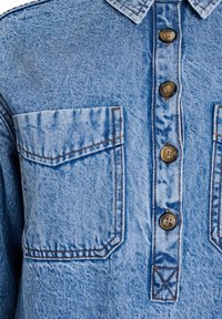 Chemise en denim bleu clair avec deux poches poitrine, boutons marron, col pointu et motifs de couture visibles. La texture est légèrement usée.