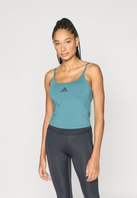 Turquoise sporttop met dunne bandjes en een zwart adidas-logo in het midden van de borst, gecombineerd met zwarte leggings. Soepele stofstructuur.
