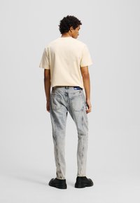 T-shirt jaune clair à manches courtes, associé à un jean bleu délavé. Le t-shirt a une coupe décontractée ; le jean présente des poches arrière et une légère tapered. Chaussures noires.